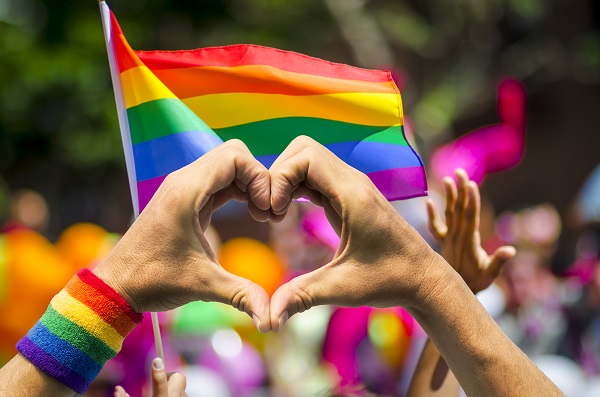 Two hands forming a heart wearing a rainbow wrist band in front of a Gay Pride flag.